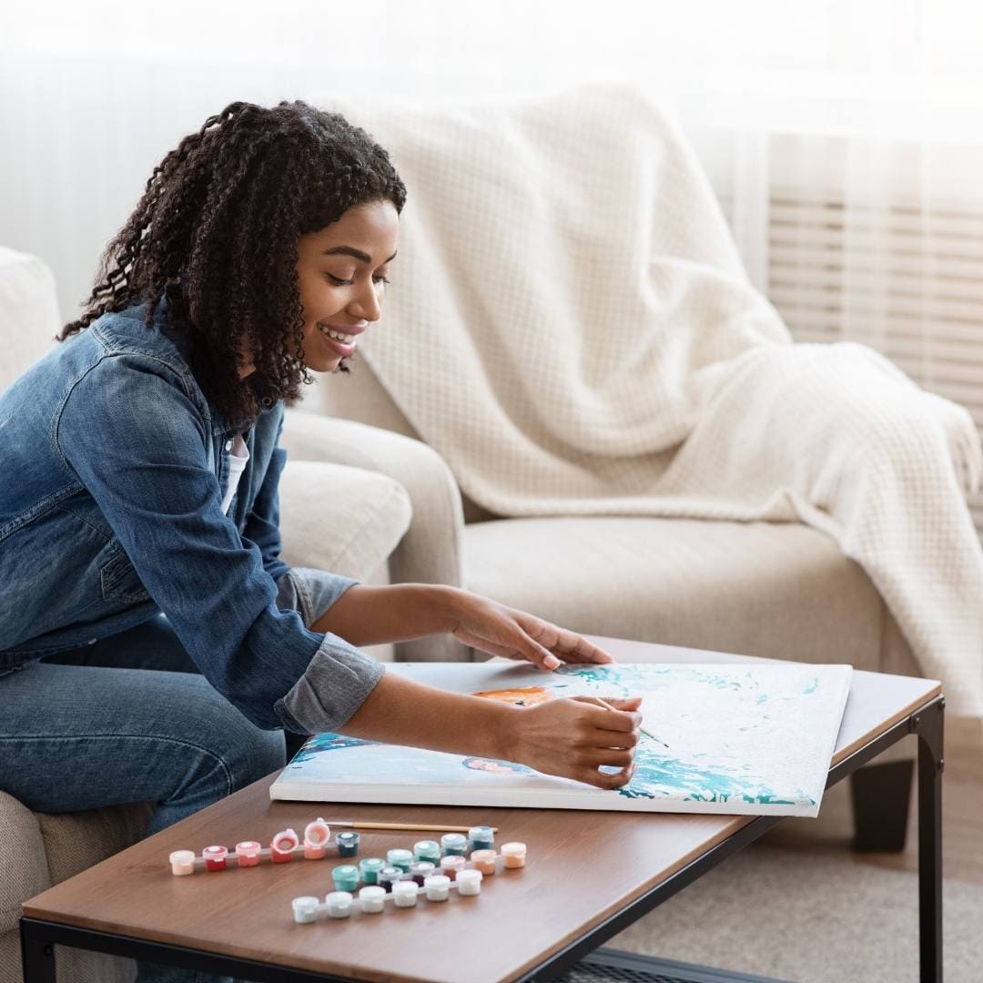 Junge Frau entspannt beim Malen nach Zahlen auf einer Leinwand – Farbsets und Pinsel auf dem Tisch – kreative Auszeit im Wohnzimmer